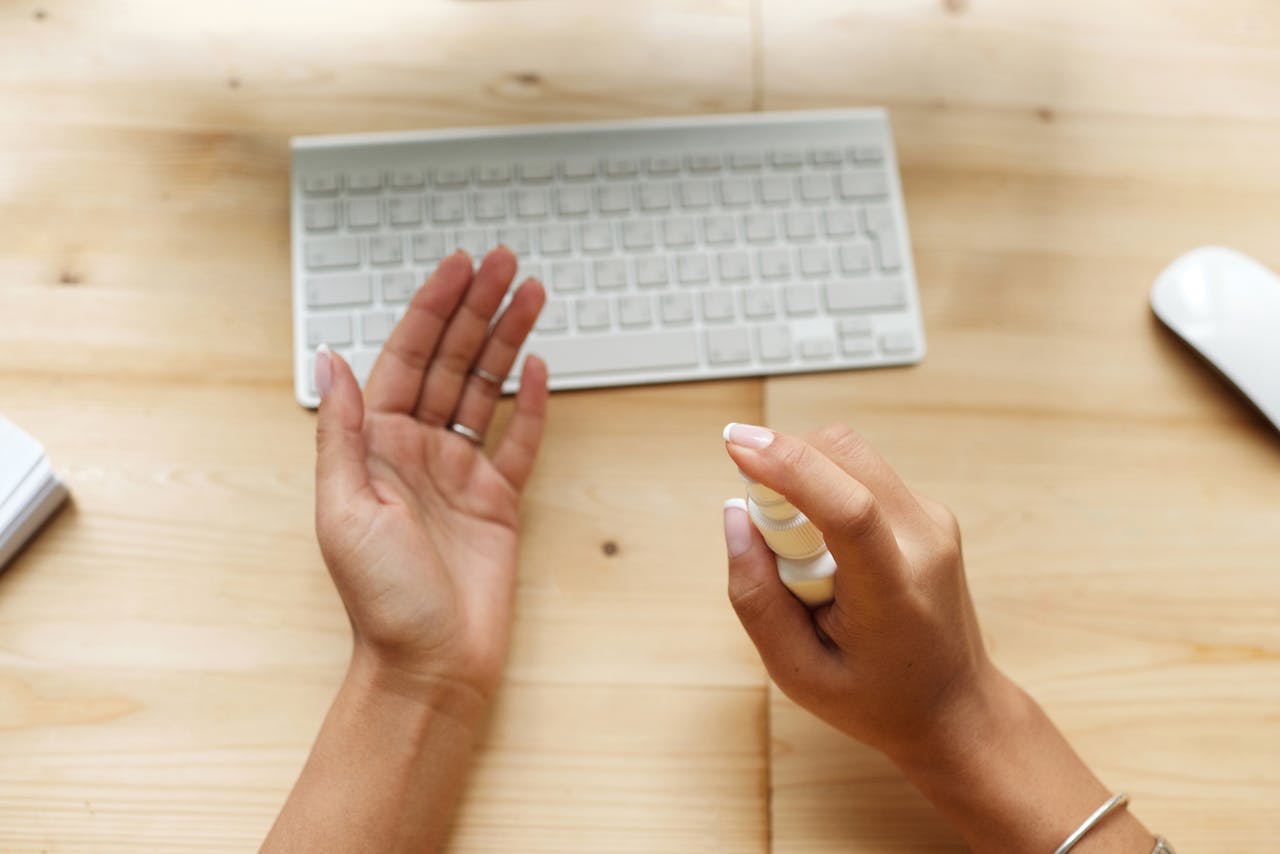 hero-img Close-up of hands using sanitizer spray near a keyboard on a wooden desk.
