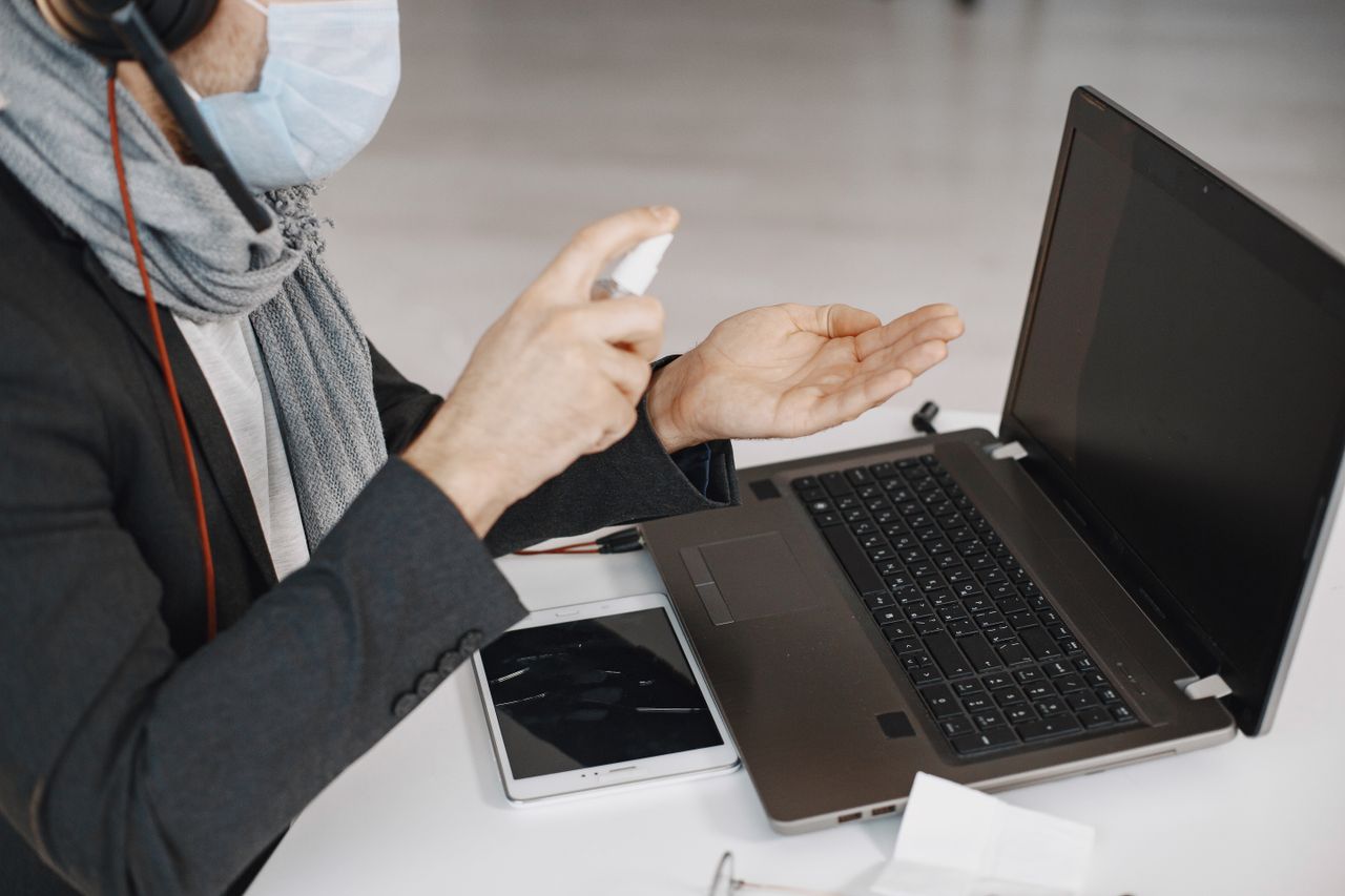 about-01 A man wearing a mask sanitizing his hands while working at a laptop indoors.